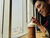 a man drinking a cup of ramen with chopsticks in front of a window